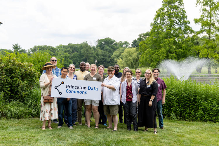 Group photo of contributor to Princeton Data Commons. Group of 16 people standing outside on the grass in front of a fountain holding a sign that reads 'Princeton Data Commons'.